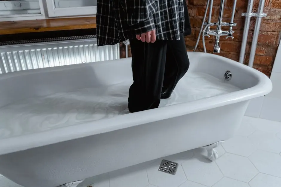 A person standing in a water-filled bathtub indoors wearing casual clothes.