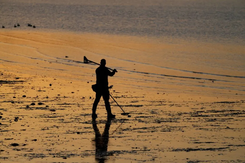Silhouette of a man using a metal detector on a beach during sunset, creating a peaceful and reflective scene.